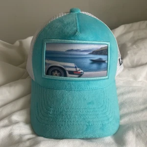 Turkos och vit keps - Snygg turkos keps med vit mesh och ett tryck av en bil och havsutsikt på framsidan. Märket LA Stilé är tryckt på insidan och sidan av kepsen. Perfekt för en solig dag! Pris kan diskuteras.