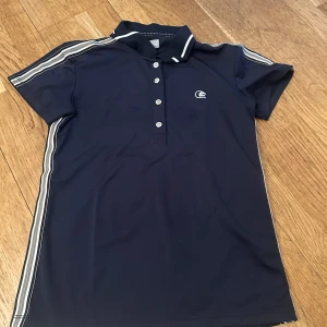 Mörkblå polotopp från Fair Play - Säljer en mörkblå polotopp från Fair Play med vita ränder längs ärmarna och knappar framtill. Toppen har korta ärmar och en klassisk krage med vita detaljer. Perfekt för en sportig look!