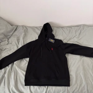 Polo Ralph Lauren  - Säljer en stilren svart hoodie från Ralph Lauren med det klassiska röda logotypbroderiet på bröstet. Hoodien har en justerbar huva med dragsko och en praktisk magficka. Perfekt för en avslappnad look.