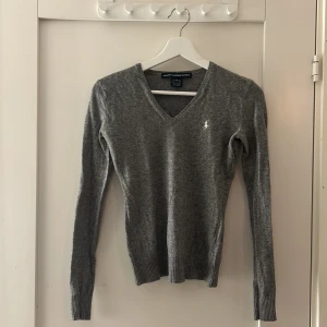 Grå v-ringad tröja från Ralph Lauren Sport - Säljer en stilren grå v-ringad tröja från Ralph Lauren Sport. Tröjan är tillverkad i en mjuk blandning av merinoull, nylon, viskos och kashmir. Den har långa ärmar och en liten broderad logotyp på bröstet. Perfekt för en elegant och bekväm look.