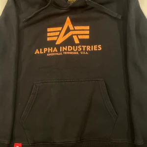 Svart hoodie från Alpha Industries - Säljer en svart hoodie från Alpha Industries med orange logotyp och text på framsidan. Tröjan har en klassisk känguruficka och justerbar huva med dragsko. Perfekt för en avslappnad stil. 250kr