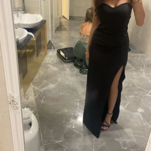 Svart axelbandslös klänning - Elegant svart axelbandslös klänning med en hög slits framtill. Perfekt för en kväll ute eller en speciell tillställning. Klänningen har en figurnära passform som framhäver silhuetten.