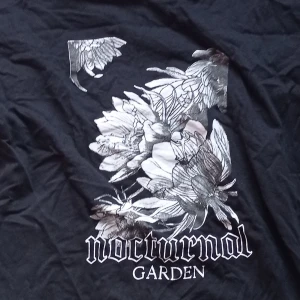 Svart t-shirt med blommigt tryck från Black Label Collection - Snygg svart t-shirt med ett stort blommigt tryck och texten 'nocturnal garden' på framsidan. Välanvänd och därför lite rynklig men det är det enda defekten som kan strykas bort