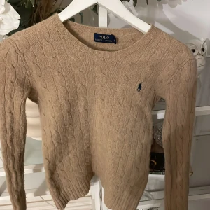 Beige stickad tröja från Polo Ralph Lauren - Säljer en snygg beige stickad tröja från Polo Ralph Lauren med kabelstickat mönster och den klassiska loggan på bröstet. Tröjan har långa ärmar och en rund halsringning. Den är skön och väldigt mjuk🥰💓lite knopprig men ingenting man tänker på😊