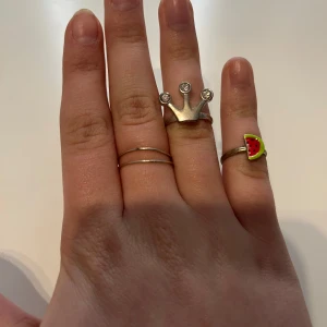 Set med tre ringar - Säljer ett set med tre unika ringar. En ring har en kronform med små stenar, en annan är enkel och tunn, och den tredje har en färgglad vattenmelon-design. Perfekt för att mixa och matcha!