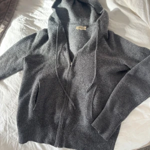 Grå hoodie från Soft Goat - Säljer en mysig grå hoodie från Soft Goat. Den har en dragkedja framtill och en justerbar huva med snören. Tröjan är tillverkad i ett mjukt material och har ribbade muddar vid ärmsluten. Perfekt för sommaren och våren
