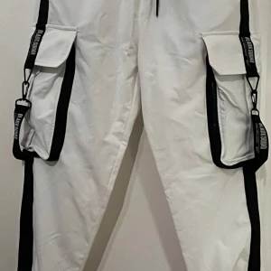 Vita cargopants från Black Squad - Snygga vita cargopants från Black Squad med svarta detaljer och stora fickor. Byxorna har justerbara remmar och en cool design. Nya men lapp saknas.