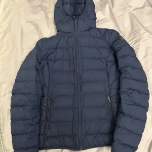 Mörkblå dunjacka från Parajumpers - Säljer en snygg mörkblå dunjacka från Parajumpers med huva och dragkedja. Jackan har en broderad logotyp på ärmen och är perfekt för kyliga dagar. Den är både stilren och funktionell pris kan sänkas vid snabb affär!