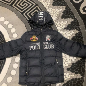 Svart dunjacka från Vinson Polo Club - Säljer en svart dunjacka från Vinson Polo Club med huva och dragkedja. Jackan har broderade detaljer på bröstet och är perfekt för kyligare väder. Den är långärmad och har en sportig look.