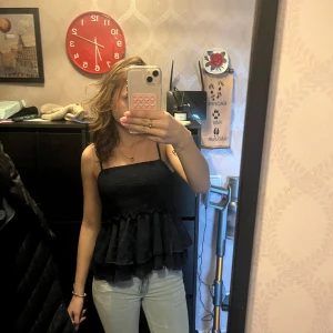 Svart axelbandslös topp - Säljer en stilren svart axelbandslös topp med volangdetaljer. Perfekt för en kväll ute eller en somrig dag. Toppen har en luftig passform och är lätt att matcha med olika outfits.