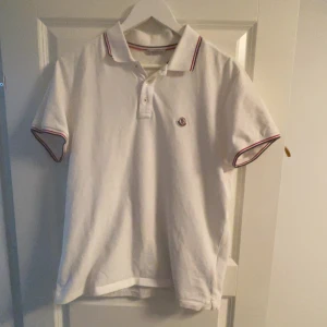 Vit pikétröja från Moncler - Säljer en stilren vit pikétröja från Moncler med korta ärmar och klassisk krage. Tröjan har röd och blå randdetalj på krage och ärmslut samt en broderad logotyp på bröstet. Perfekt för en avslappnad men ändå snygg look. Tröjan har en fläck bak på ryggen som syns på bild 3 men den ger ett bra helhetsintryck.