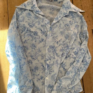 Blåvit blommig skjorta från Otex - Säljer en stilren blåvit skjorta från Otex med ett vackert blommigt mönster. Skjortan har långa ärmar och 70 tals krage. Är antagligen gjord runt 70 talet. Storleken uppskattas vara runt S
