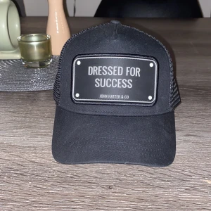 Svart keps från John Hatter & Co - Snygg svart keps med texten 'Dressed for Success' på framsidan. Kepsen har ett nätmaterial på baksidan för extra ventilation och en klassisk böjd skärm. Perfekt för en avslappnad stil. Nytt skicka. Aldrig använd 