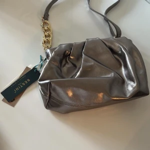 Silver axelväska från Bestini - Snygg silverfärgad axelväska från Bestini med en glansig finish. Väskan har en kedjedetalj i guld på axelremmen som ger en lyxig touch. Perfekt för att lyfta vilken outfit som helst.
