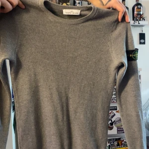 Grå stickad tröja från Stone Island - Säljer en stilren grå stickad tröja från Stone Island. Tröjan har en ribbad struktur och loggan på ärmen. Är en storlek S men passar även som en M. 