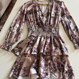 Blommig klänning med volanger - Säljer en vacker blommig klänning med volanger och v-ringning. Klänningen har långa ärmar och ett elastiskt midjeband som ger en smickrande passform. Perfekt för vår och sommar!