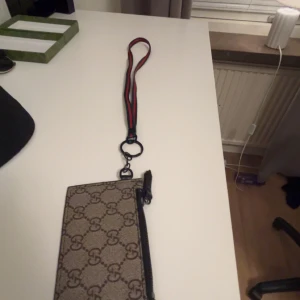 Mönstrad plånbok med rem - Säljer en stilren plånbok med ett klassiskt mönster och en praktisk dragkedja. Plånboken har en avtagbar rem i rött och grönt, perfekt för att bära runt halsen eller i handen. Kommer i originalförpackning. (Med box)