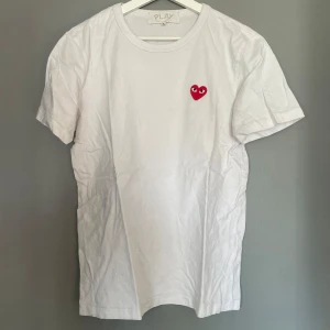 Vit t-shirt med rött hjärta från Comme des Garçons - Säljer en stilren vit t-shirt från Comme des Garçons med ett ikoniskt rött hjärta på bröstet. Perfekt för en casual look. T-shirten har en normal passform och är kortärmad.