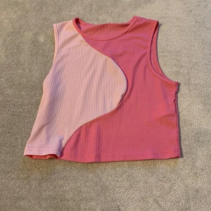 Rosa och ljusrosa ribbad linnetopp - Säljer en cool och trendig rosa och ljusrosa ribbad linnetopp. Toppen har ett asymmetriskt mönster framtill och är ärmlös, vilket gör den perfekt för varma dagar. Den är croppad och passar bra till både jeans och kjol. Perfekt för sommaren! 🌸