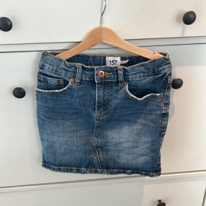 Blå jeanskjol från 157 - Snygg blå jeanskjol från 157 med klassisk femficksdesign och knappstängning fram. Perfekt för en avslappnad stil. Passar bra till både sneakers och sandaler.