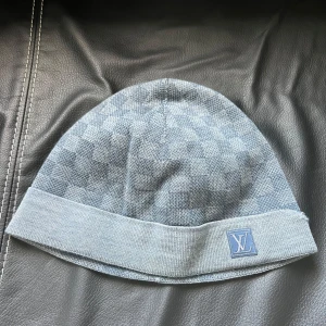 Ljusblå rutig mössa från Louis Vuitton - Snygg ljusblå mössa från Louis Vuitton med ett diskret rutmönster. Mössan har en uppvikt kant och en liten logotyp framtill. Perfekt för att hålla stilen i kallare väder. Mössan har en mindre defekt som knappt syns när man bär mössan. Denna mössa från Louis Vuitton tillverkas inte längre därav priset även fast den har en defekt.