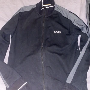 Svarta mjukisbyxor från BOSS - Snygga svarta mjukisbyxor från BOSS med grå sidopaneler och snörning i midjan. Perfekta för en avslappnad stil med en sportig touch. BOSS-loggan är diskret placerad på benet. Även en fin boss kofta. Dressen är väldigt bekväm och kan användas ungefär när du känner för det. Koftan är M, byxorna L