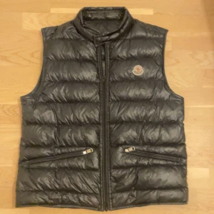 Monclear väst gui - Äkta Snygg svart monclear dunväst i storlek 3 med dragkedja som är bytt (original blixtlås följer med). Västen har två praktiska fickor med dragkedjor framtill och en diskret logga på bröstet. Perfekt för kyligare dagar. Pris går att diskutera
