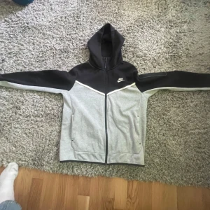 Grå och svart nike tech fleece - Säljer en snygg grå och svart hoodie från Nike med dragkedja och huva. Perfekt för en sportig look eller avslappnade dagar. Den har långa ärmar och en klassisk Nike-logga på bröstet.