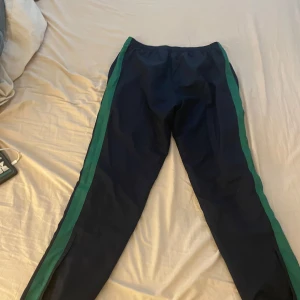 Blå träningsbyxor från Lacoste - Snygga blå träningsbyxor från Lacoste med gröna ränder längs sidorna. Byxorna har en elastisk midja och en dragkedja vid ben änden. Perfekta för en sportig och klassisk look. Nypris ~1200