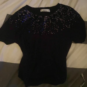 Svart kortärmad topp från Zara med paljetter - Snygg svart topp från Zara med korta ärmar och glittrande paljetter över axlarna och bröstet. Perfekt för en kväll ute eller en festlig tillställning. Toppen har en rund halsringning och en figurnära passform.