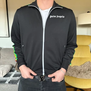 Svart Palm Angels kofta  - Säljer min killes äkt svarta Palm Angels kofta med vita dragkedjor och färgglada ränder i grönt, gult och rött längs ärmarna. L men passar även som M