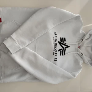 Vit hoodie från Alpha Industries - Säljer en stilren vit hoodie från Alpha Industries med svart logotyptryck på framsidan. Hoodien har en klassisk design med känguruficka och ribbade muddar. Perfekt för en avslappnad stil.