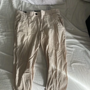 Beige chinos från Jack & Jones - Snygga beige chinos från Jack & Jones med normal passform. Byxorna har en klassisk design med knappar och dragkedja framtill. Perfekta för en stilren look.