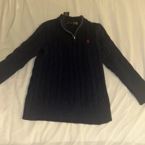 Mörkblå stickad tröja från Polo Ralph Lauren - Säljer en snygg mörkblå stickad tröja från Polo Ralph Lauren med dragkedja och det klassiska röda logotypbroderiet på bröstet. Tröjan har en ribbad krage och långa ärmar, perfekt för kyligare dagar. Passar både till jeans och chinos!