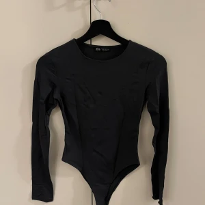 Mörkgrå långärmad bodysuit från Zara - Snygg svart långärmad bodysuit från Zara. Perfekt för en stilren look med sin enkla design och åtsittande passform. Tillverkad i ett mjukt och stretchigt material för extra komfort.