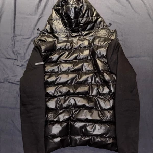 Svart dunväst från Koroshi - Snygg svart dunväst från Koroshi med huva och dragkedja. Västen har en glansig finish och är perfekt för kyligare dagar. Den har en modern look med svarta ärmar och en quiltad design.