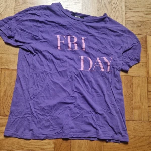 Lila t-shirt från Bik Bok - Säljer en lila t-shirt från Bik Bok med texten 'FRIDAY' i rosa på framsidan. Strl M, skönt material. Perfekt till träning eller färgklick i vardagen. Katt i hemmet. 