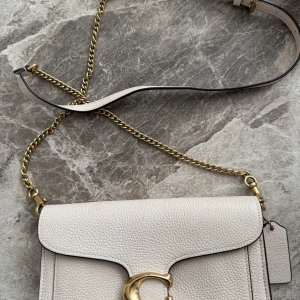 Beige axelväska med gulddetaljer - Elegant beige axelväska med guldfärgad kedja och spänne. Väskan har en stilren design med en praktisk dragkedja inuti. Perfekt för att bära dina nödvändigheter med stil. Använd endast en gång, priset kan diskuteras.