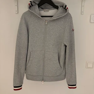 Grå hoodie från Moncler - Säljer en stilren grå hoodie från Moncler med dragkedja och huva. Tröjan har ribbade muddar och Moncler-logga på ärmen. Perfekt för en avslappnad stil.