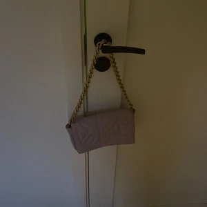 Lila axelväska med kedja - Snygg lila axelväska med quiltad design och guldfärgad kedja som axelrem. Perfekt för att ge en touch av stil till din outfit. Väskan har en praktisk dragkedja inuti för säker förvaring.