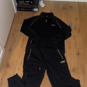 Svart träningsset från Boss - Säljer ett stilrent svart träningsset från Boss med dragkedja och gulddetaljer. Setet består av en långärmad tröja och matchande byxor, perfekt för en sportig look. Tröjan har en dragkedja framtill och byxorna har snörning i midjan.