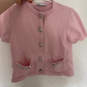 Rosa stickad tröja från ZARA - Söt rosa stickad tröja från ZARA med korta ärmar och dekorativa knappar. Den har två små fickor fram med fina detaljer. Perfekt för en chic och avslappnad look. 200kr
