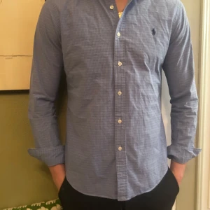 Blå rutig skjorta från Ralph Lauren - Snygg blå rutig skjorta från Ralph Lauren med klassisk krage och knappar framtill. Skjortan har långa ärmar och en broderad logga på bröstet. Perfekt för en stilren look.