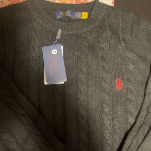 Polo Ralph Lauren stickad tröja  - Säljer en stilren grå stickad tröja från Polo Ralph Lauren med klassiskt flätmönster. Tröjan har en broderad röd logotyp på bröstet och rund halsringning. Perfekt för kyliga dagar.