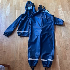 Blå regnjacka med hängselbyxor - Säljer en blå regnjacka med matchande hängselbyxor. Jackan har en huva och långa ärmar med elastiska muddar. Byxorna har justerbara hängslen och elastiska benslut. Perfekt för regniga dagar!