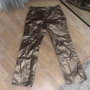 Guldiga byxor med glansig finish - Snygga guldiga byxor med en glansig finish. De har en klassisk femficksdesign och stängs med dragkedja och knapp. Perfekta för att sticka ut med en unik stil.