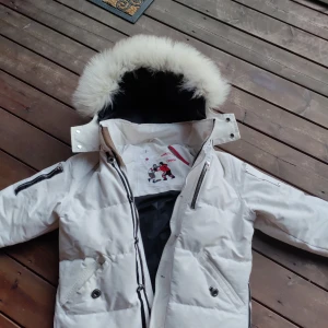Vit dunjacka från Moose Knuckles - Säljer en snygg vit dunjacka från Moose Knuckles med pälskantad huva. Jackan har dragkedja och knappar framtill samt broderad detalj på insidan