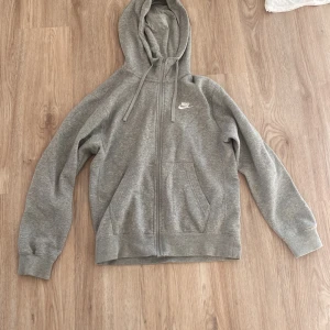 Grå hoodie från Nike - Säljer en grå hoodie från Nike med dragkedja och huva. Tröjan har ett broderat Nike-märke på bröstet och är fodrad med mjukt material för extra komfort. Tröjan är storlek XS och är nyskick. Hör av dig vid funderingar!🙌