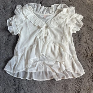 Vit volangblus från Nelly - Supersöt vit blus från Nelly med volanger och v-ringning. Blusen har korta puffärmar och är gjord i ett tunt, luftigt material som ger en romantisk vibe. Perfekt för dig som gillar lager på lager och detaljer som sticker ut.