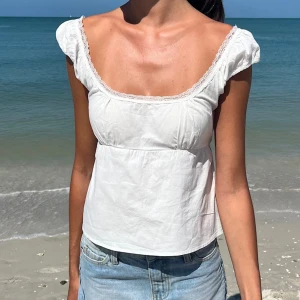 Brandy Melville Vit blus med spets och knyt - Supersöt vit blus med korta puffärmar och spetskant runt den vida urringningen. Blusen har knyt i ryggen för en fin passform och är gjord i ett lätt bomullstyg. Perfekt för varma dagar och ger en romantisk vibe till din outfit.
